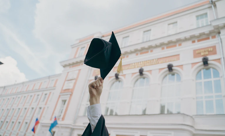 Hand holding a graduation cap before a university building, representing a bachelor's degree